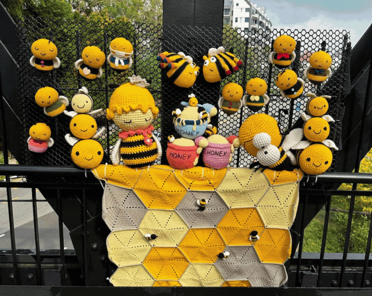 Crochet amigurumi bees and honey pots arranged on a hexagonal honeycomb blanket, displayed on a black metal bridge railing outdoors.